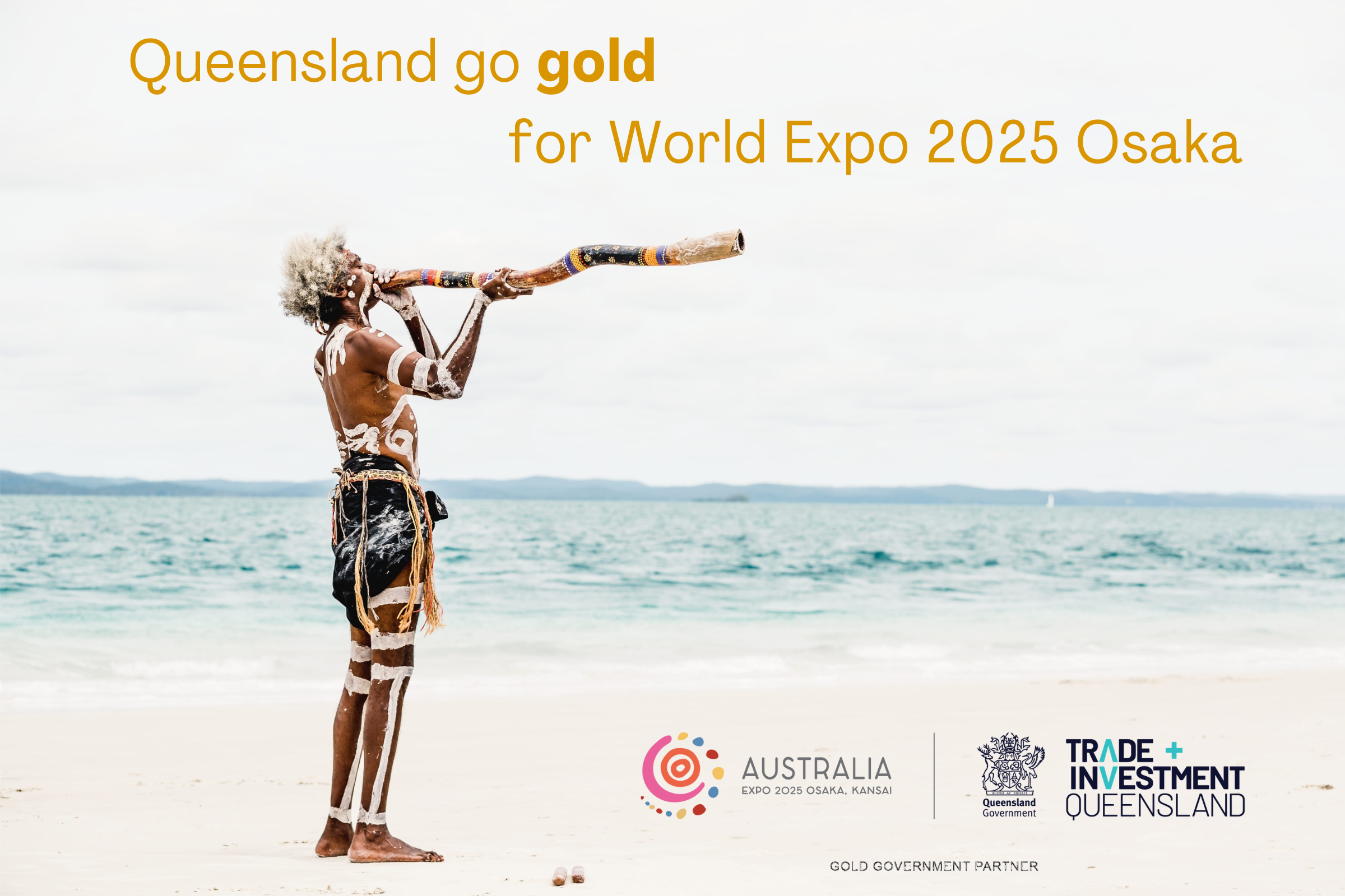 A First Nations man in traditional dress stands on the beach of K'Gari Island, Australia playing the didgeridoo. The blue ocean and clear sky are in the background.